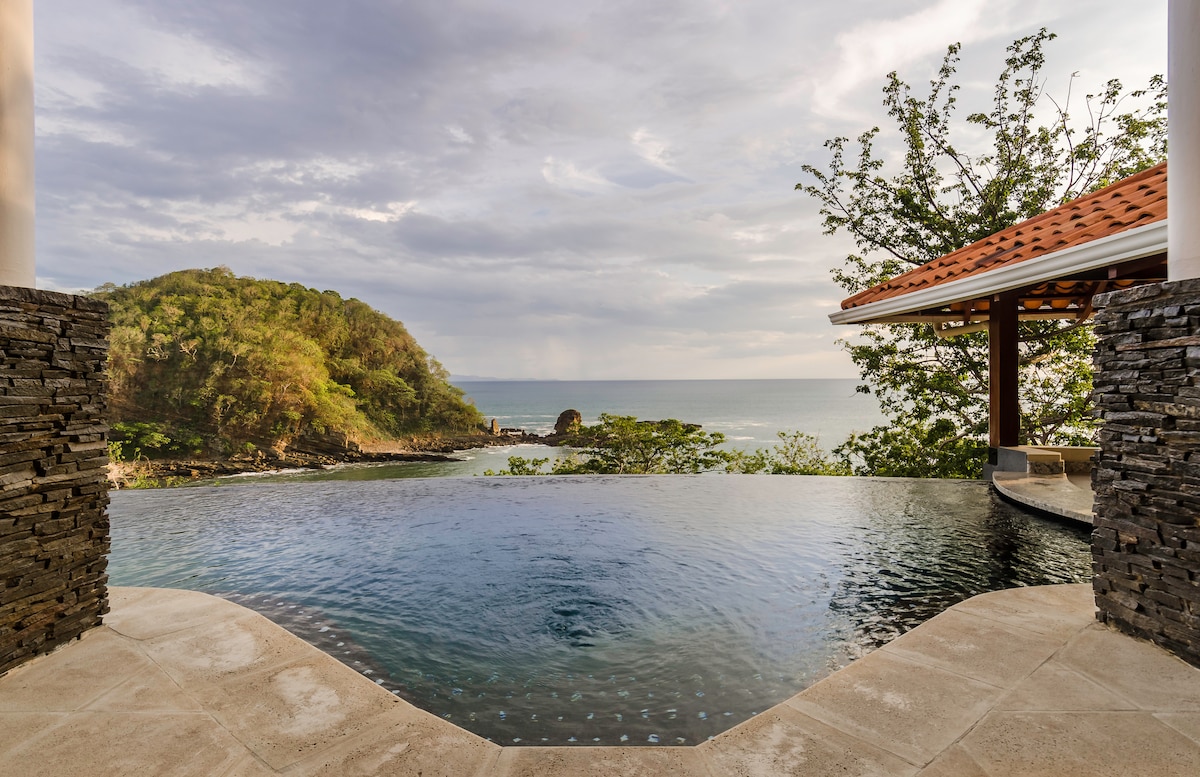 An infinity pool is visible at the edge of a private patio, seamlessly blending into ocean views. Lush green hills frame the backdrop, while a covered seating area is situated to the right, providing shade and a relaxing atmosphere.