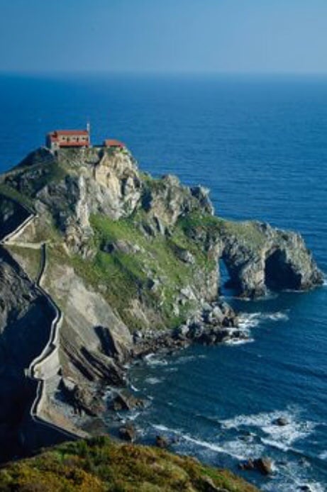 A coastal landscape features a rugged cliff adorned with lush greenery. Atop the cliff, a red-roofed structure is visible, surrounded by a serene blue sea. The path leading to the building winds along the shoreline, emphasizing the dramatic natural beauty of the area.