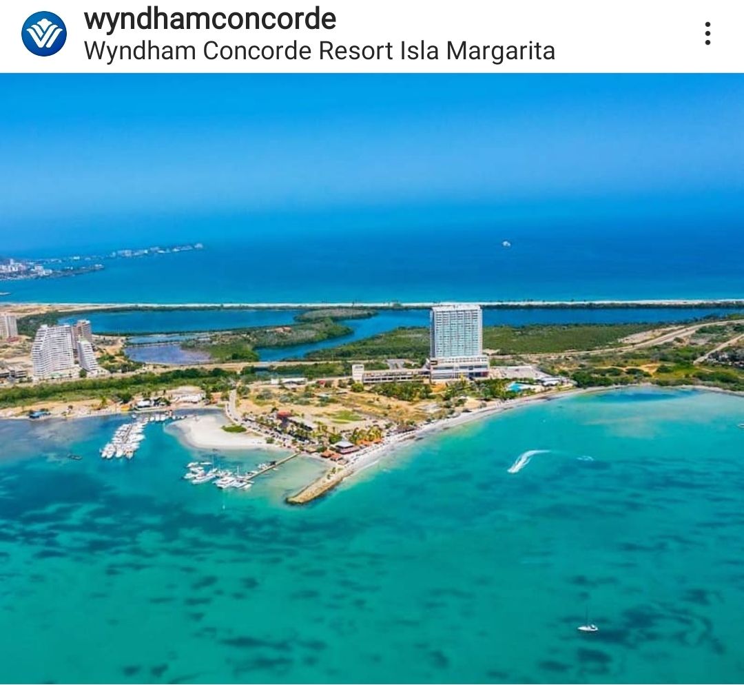 Aerial view of the Wyndham Concorde Resort Isla Margarita is displayed, showcasing the vibrant turquoise waters and sandy shores. The hotel building is positioned against a backdrop of lush greenery and the coastline, with a glimpse of the marina and boats visible along the water's edge.