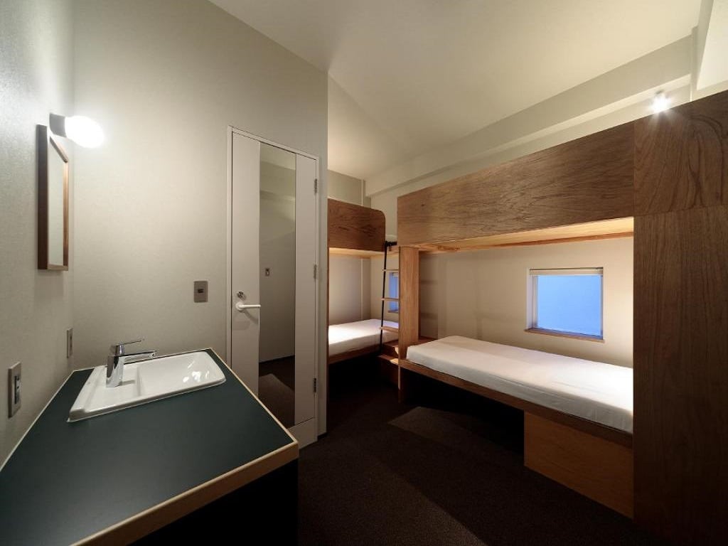 The room features two wooden bunk beds with built-in shelving and a window allowing natural light. A sink area is positioned next to the entrance. The neutral-colored walls and floor carpet provide a calm environment.