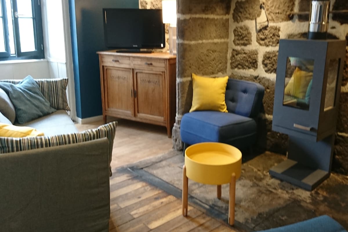 A cozy living area features a mix of modern and rustic design elements. A blue armchair and striped sofa provide seating options around a small yellow coffee table. A traditional stone wall adds character, while a television and lamp offer contemporary conveniences.