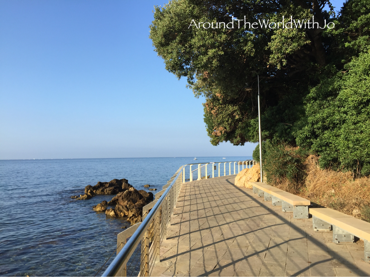 A scenic waterfront promenade is shown, lined with a white railing and bordered by lush greenery. The calm sea gently laps at the rocky shore, while the clear blue sky stretches overhead, creating a peaceful coastal view.