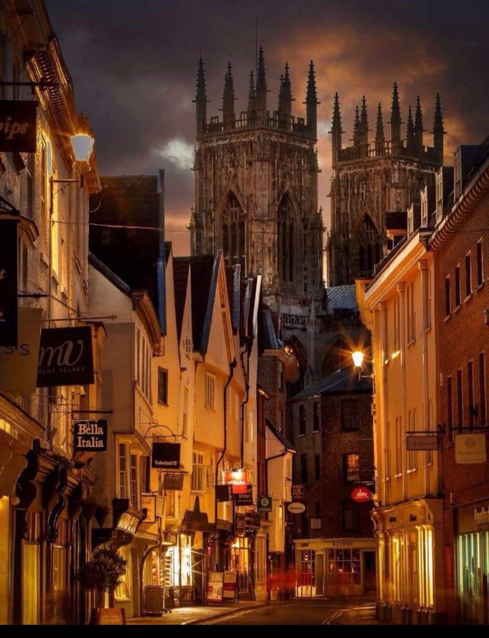 A narrow street lined with charming buildings reveals an evening view of towering cathedral spires against a dramatic sky. Warm shop lights illuminate the cobblestone pathway, inviting exploration of nearby restaurants and cafes.