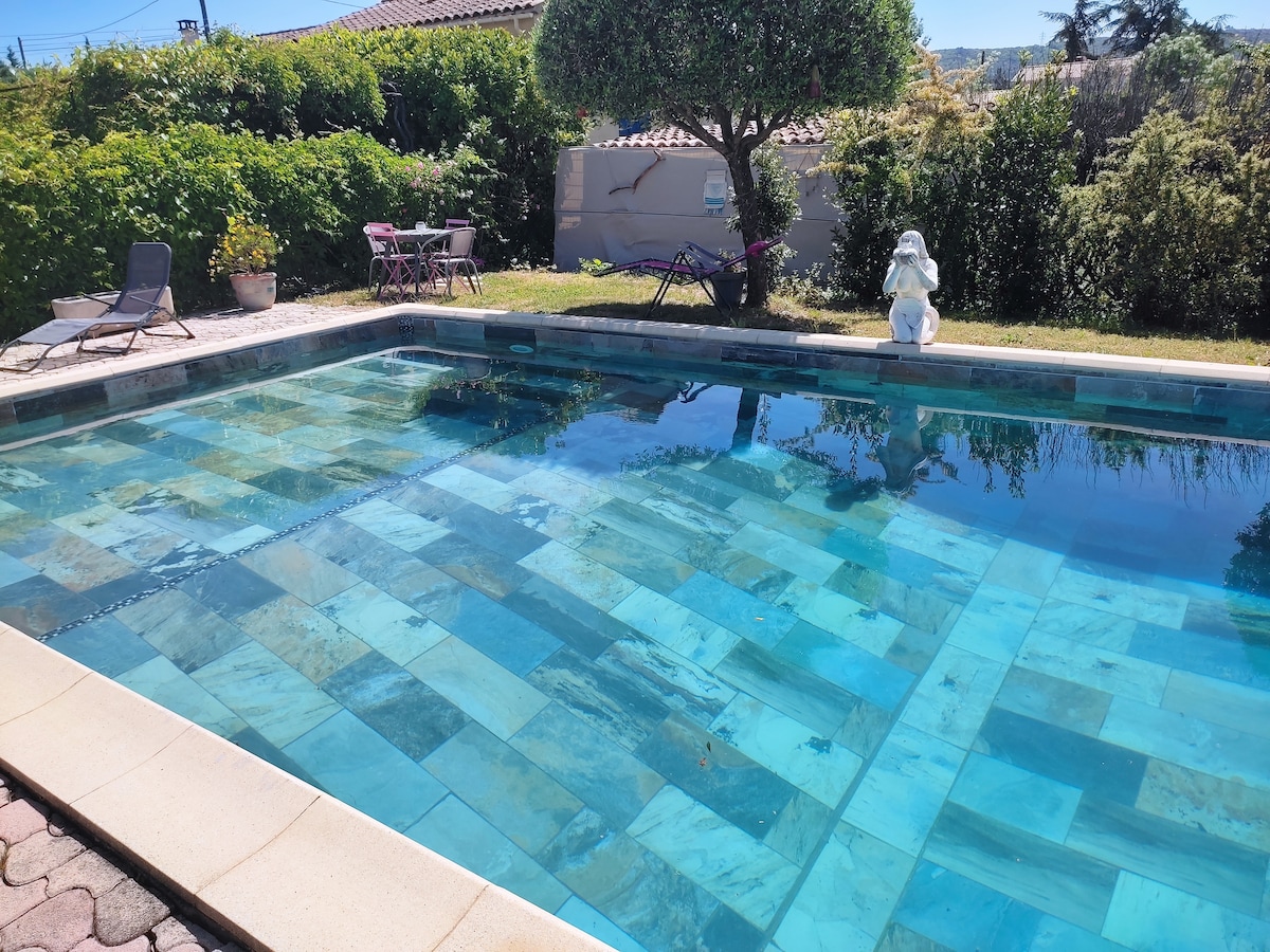 A rectangular swimming pool features a patterned stone deck and vibrant blue tiles. Sun loungers are positioned nearby on a grassy area, surrounded by shrubs and trees. A statue adds a decorative touch to the poolside, providing a tranquil setting for relaxation.