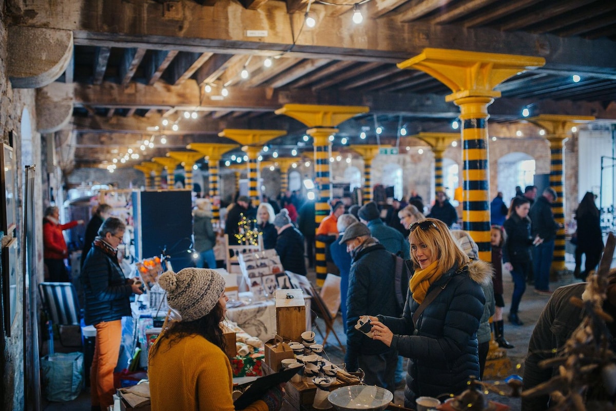 An indoor market scene features a lively gathering of people browsing various stalls under exposed beams and yellow-striped columns. Soft lighting from hanging bulbs creates a warm atmosphere, while displayed items range from crafts to food, drawing attention throughout the vibrant space.