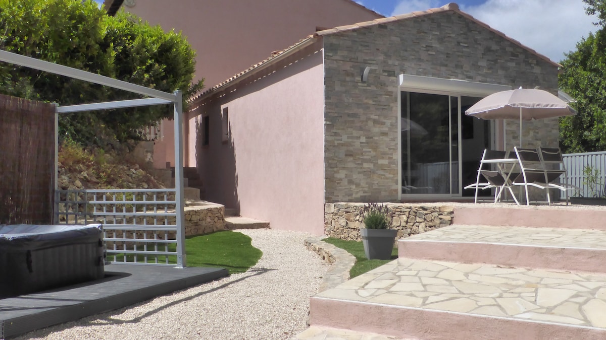 The exterior of the accommodation showcases a charming stone and pink structure surrounded by manicured greenery. A gravel pathway leads to a spacious terrace with outdoor seating and an umbrella. A hot tub is positioned to the left, enhancing the serene outdoor atmosphere.