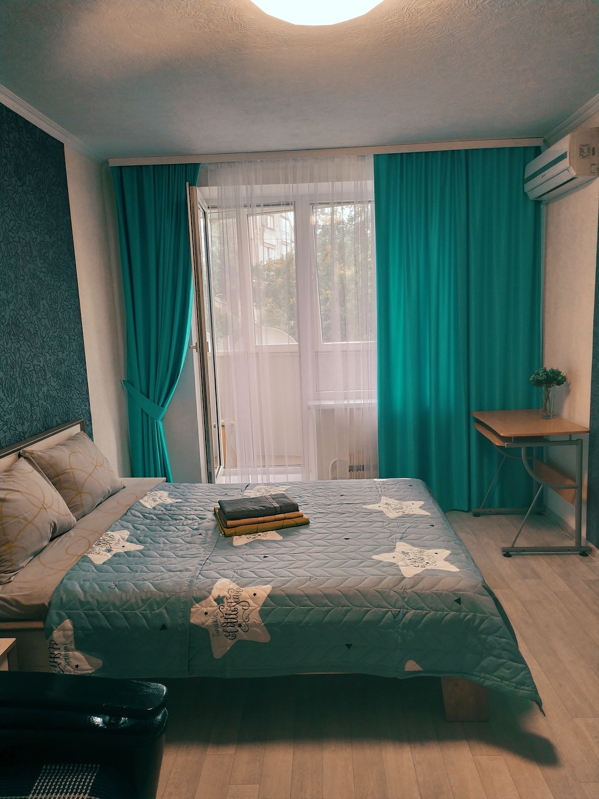 A bright sleeping area features a spacious bed with a patterned quilt draped over it. A small workspace with a chair is positioned nearby. Floor-to-ceiling curtains in turquoise frame a large window that allows natural light to enter, enhancing the serene environment.