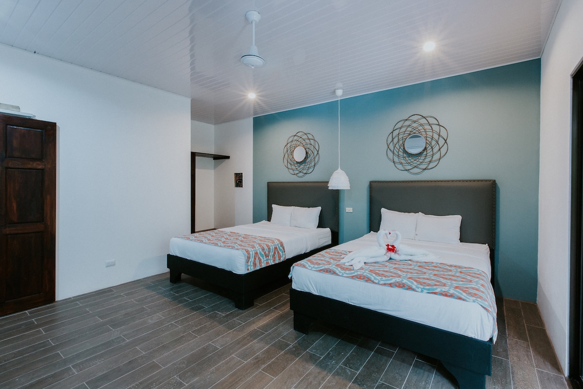 A spacious room features two large beds with neutral bedding and colorful decorative throws. Geometric wall art is positioned above each bed, complemented by stylish light fixtures. The room is filled with natural light from overhead lights, while wooden accents add warmth to the atmosphere.
