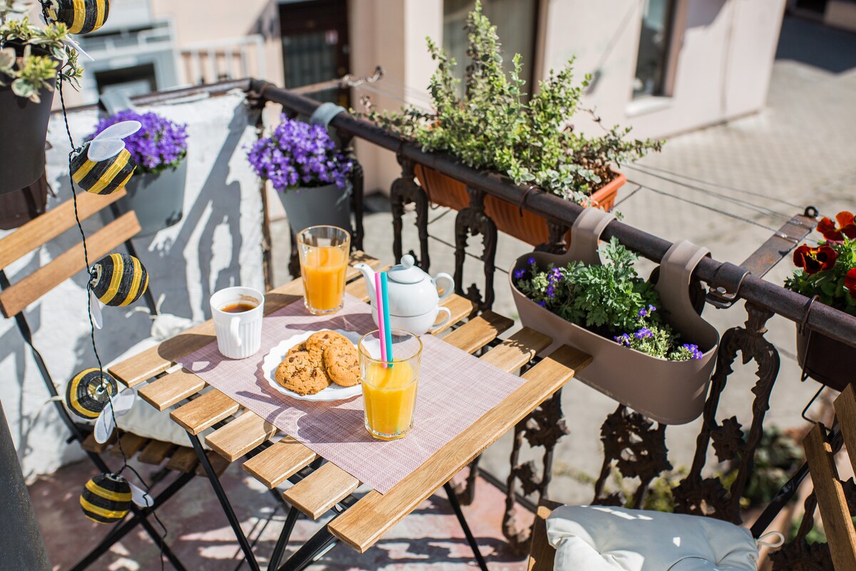 A small balcony displays a wooden table set with two glasses of juice, a cup of coffee, and a plate of cookies. Colorful flower planters hang above, adding vibrancy. The surrounding area is illuminated by daylight, creating a cheerful atmosphere for outdoor relaxation.