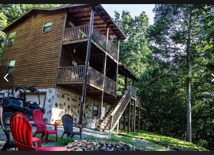 The cabin showcases a three-level structure with natural cedar siding. Multiple porches, framed by wooden railings, provide views of the surrounding greenery. Vibrant red chairs are arranged around a stone fire pit, enhancing the outdoor space. An all-terrain vehicle is parked nearby.
