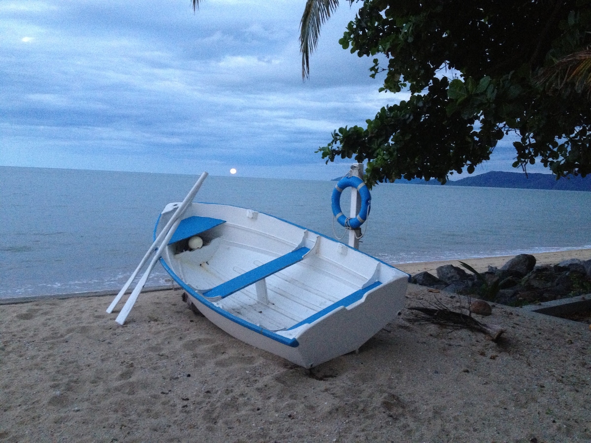 A small white boat with blue accents rests on a sandy beach, partially illuminated by a setting sun in the distance. Gentle waves lap at the shore, and palm leaves provide a touch of greenery against the sky with scattered clouds.