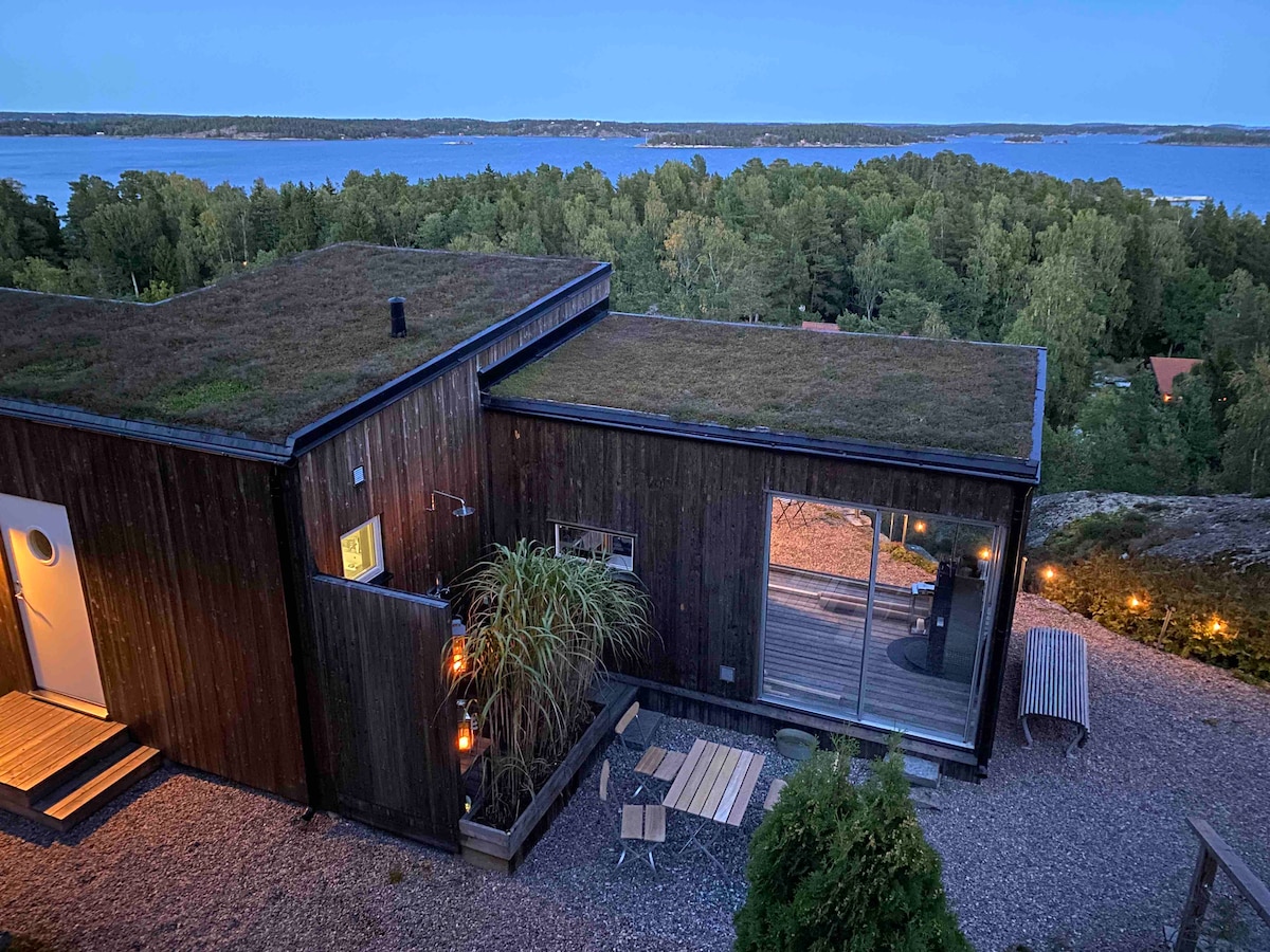 The house, designed with modern aesthetics, features a green roof and large windows reflecting the evening light. A gravel pathway leads to the entrance, while outdoor seating and lanterns create a serene ambiance. The backdrop showcases a scenic view of the water and surrounding forest.
