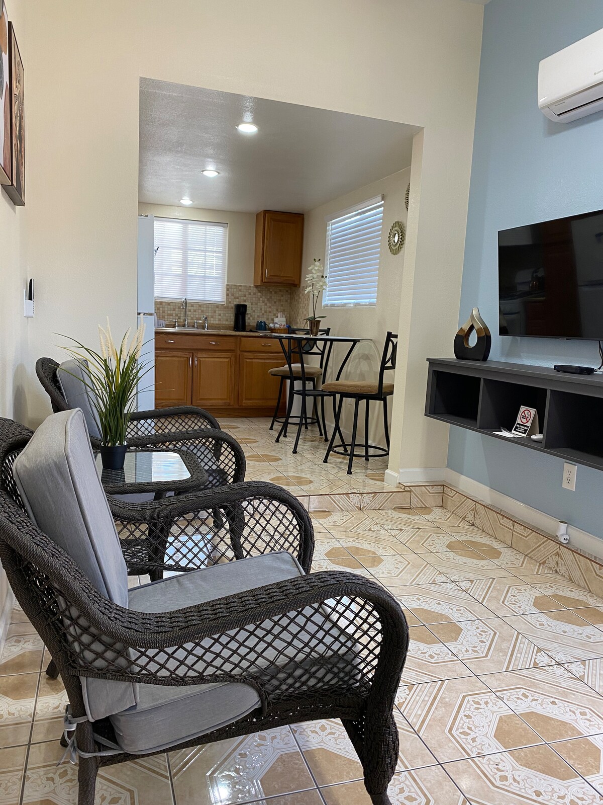 A cozy living area features comfortable wicker chairs with soft cushions, positioned on patterned tile flooring. The room opens into a well-equipped kitchen, visible in the background, with wooden cabinets and a dining table for two. Natural light brightens the space through a nearby window.