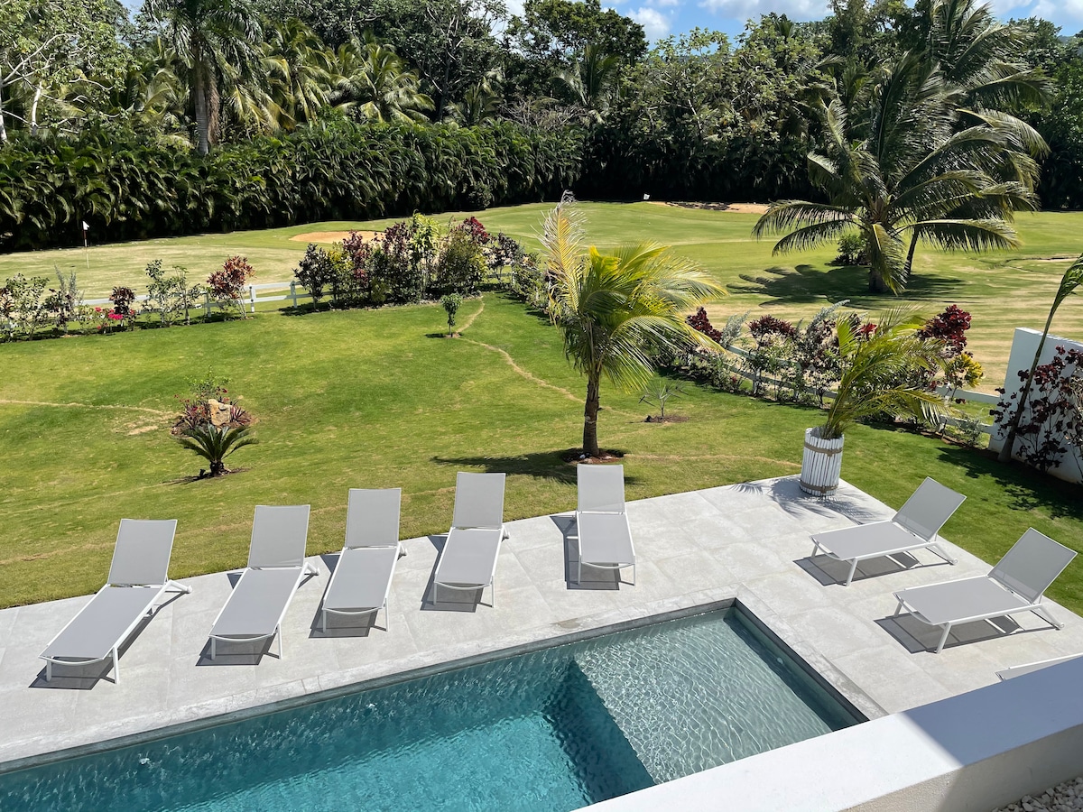 An outdoor area features a rectangular pool surrounded by a spacious patio. Eight lounge chairs are arranged neatly beside the pool, offering a relaxing space. Lush greenery and landscaped gardens provide a serene backdrop, enhancing the tranquil atmosphere.