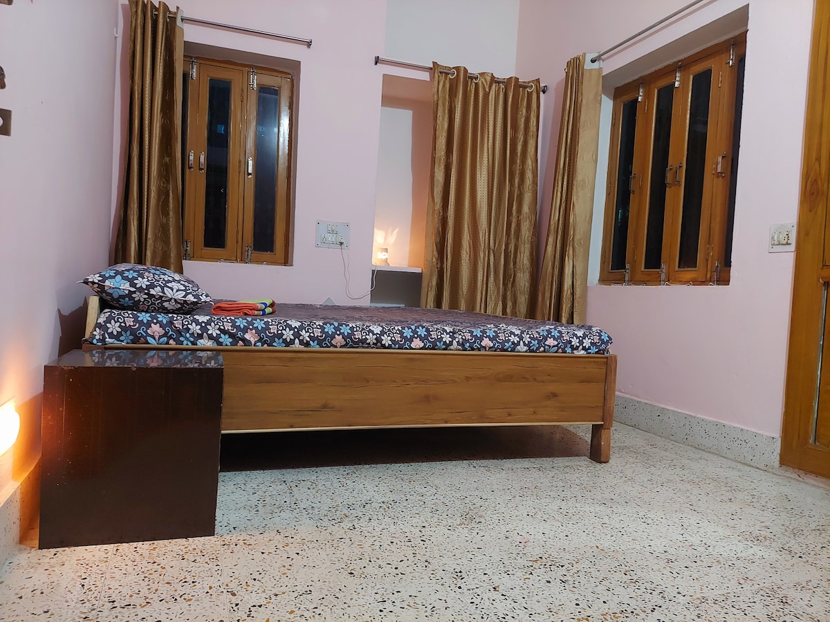 A spacious bedroom is depicted with a wooden bed featuring a floral-patterned mattress. Two windows allow natural light to enter, framed by beige curtains. A bedside table is positioned next to the bed, complementing the room's contemporary design and neutral color scheme.