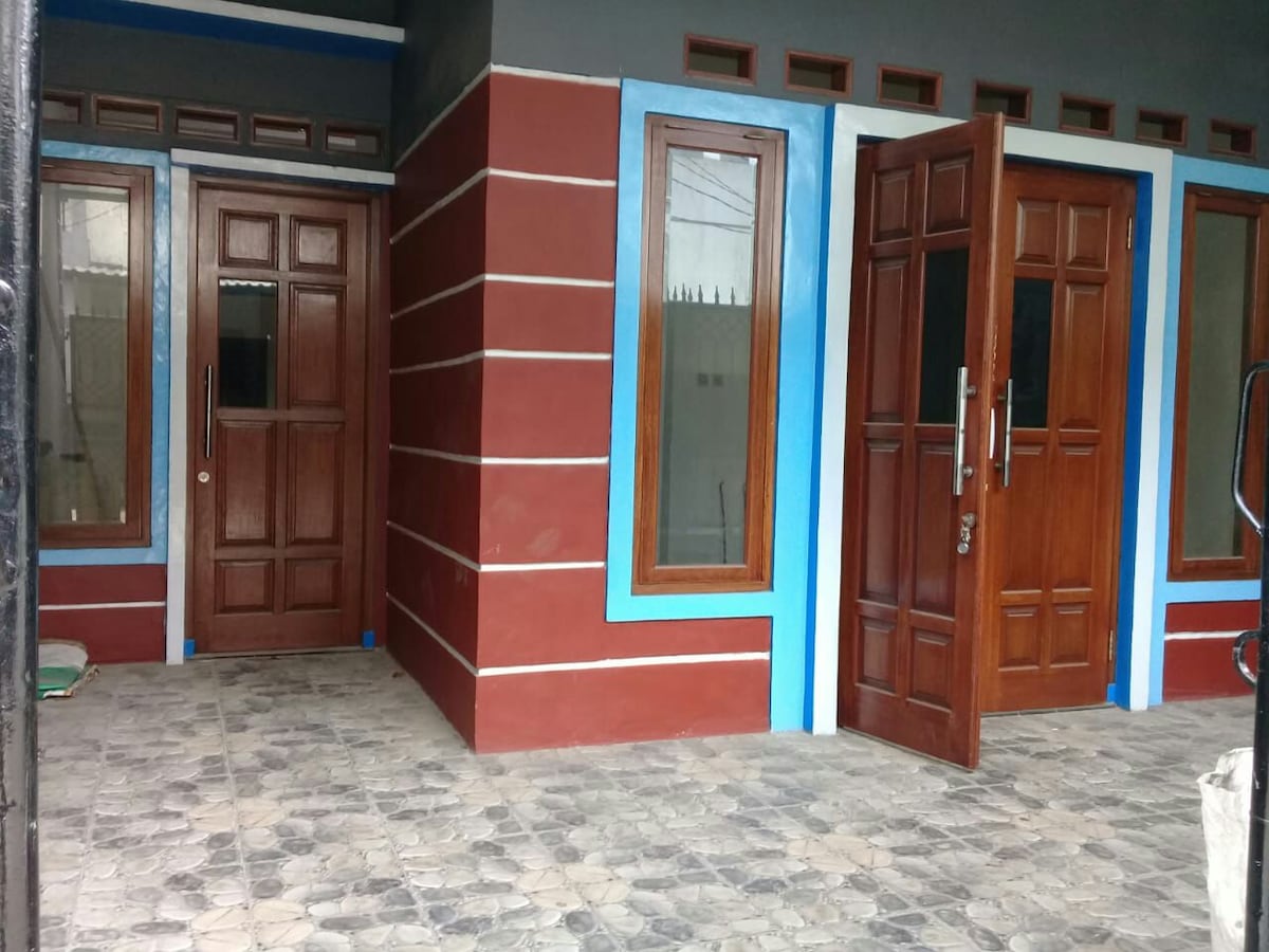 The exterior of the house features a vibrant color scheme with red and blue accents. Two wooden doors, equipped with glass panels, provide entry points. Symmetrically placed windows allow natural light to enter, and the patterned stone flooring adds texture to the space.