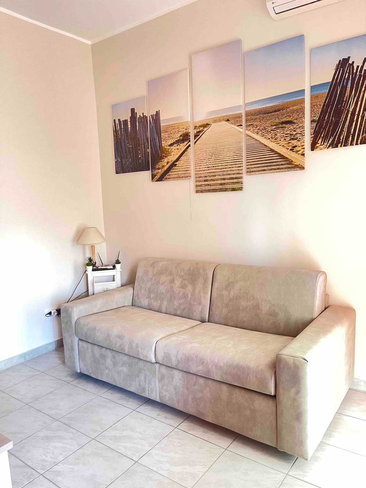 A light-colored, comfortable sofa bed is positioned against a wall adorned with a series of framed beach-themed artwork. Soft natural light enters from the nearby window, highlighting the neutral tones of the tiled floor and the furnishings.