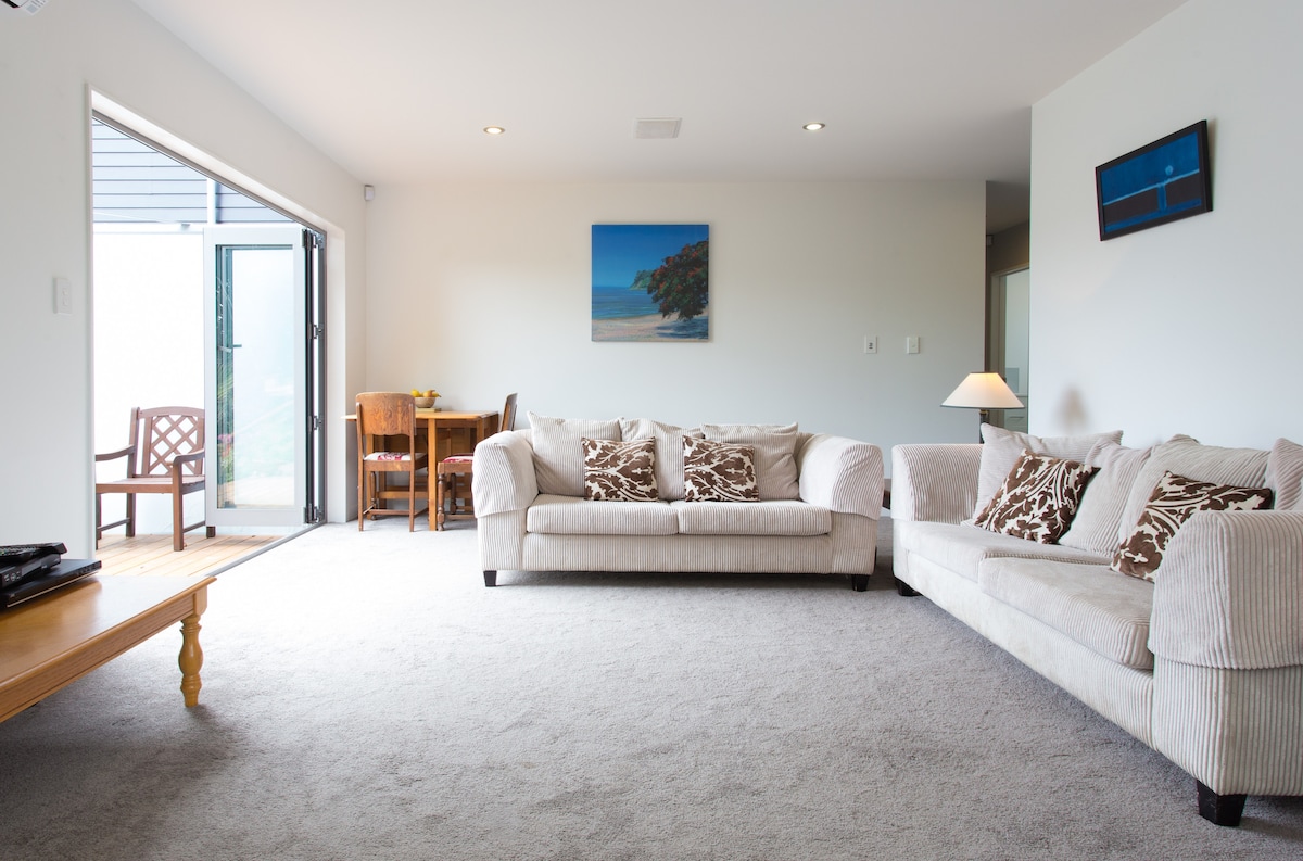 A spacious lounge area features two light-colored sofas with decorative cushions, complemented by a wooden coffee table. A small dining table with chairs is visible near the sliding glass doors that open to a patio. Natural light brightens the room.