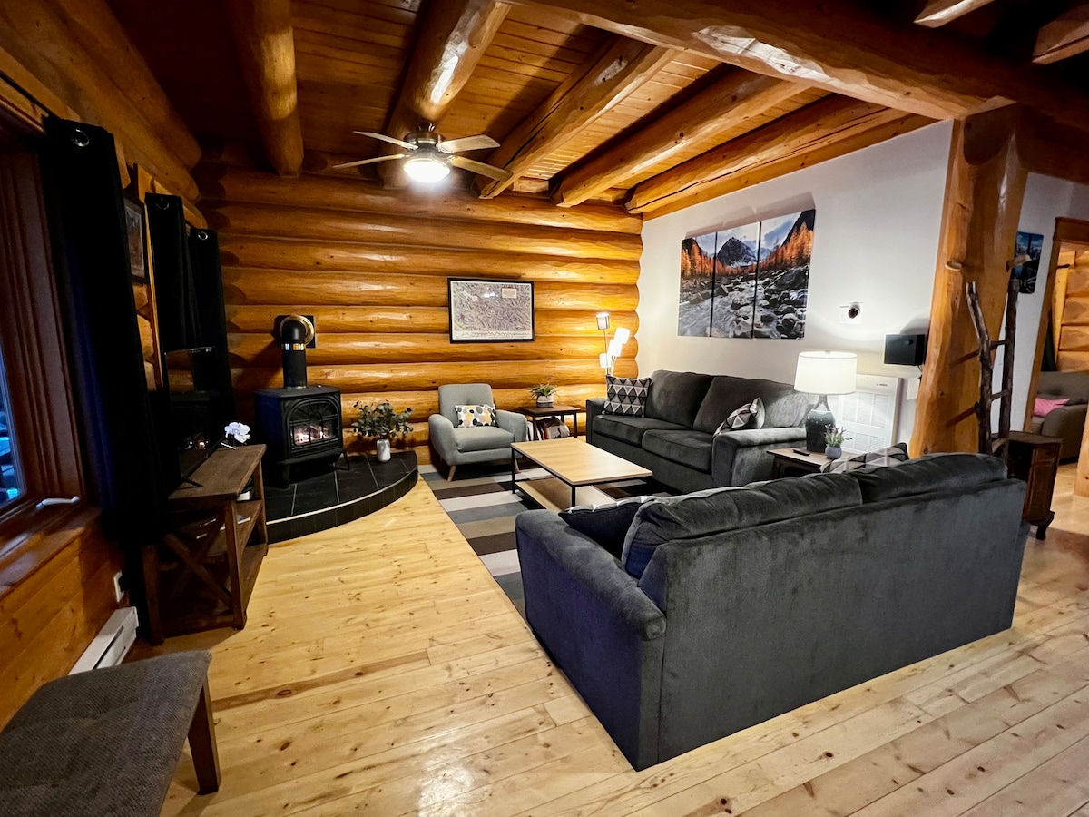 A spacious living area features log cabin walls and a warm wooden floor. Comfortable seating options include a gray sofa and armchair. A cozy fireplace is positioned against the wall, complemented by decorative artwork above. Soft lighting creates an inviting ambiance throughout the space.