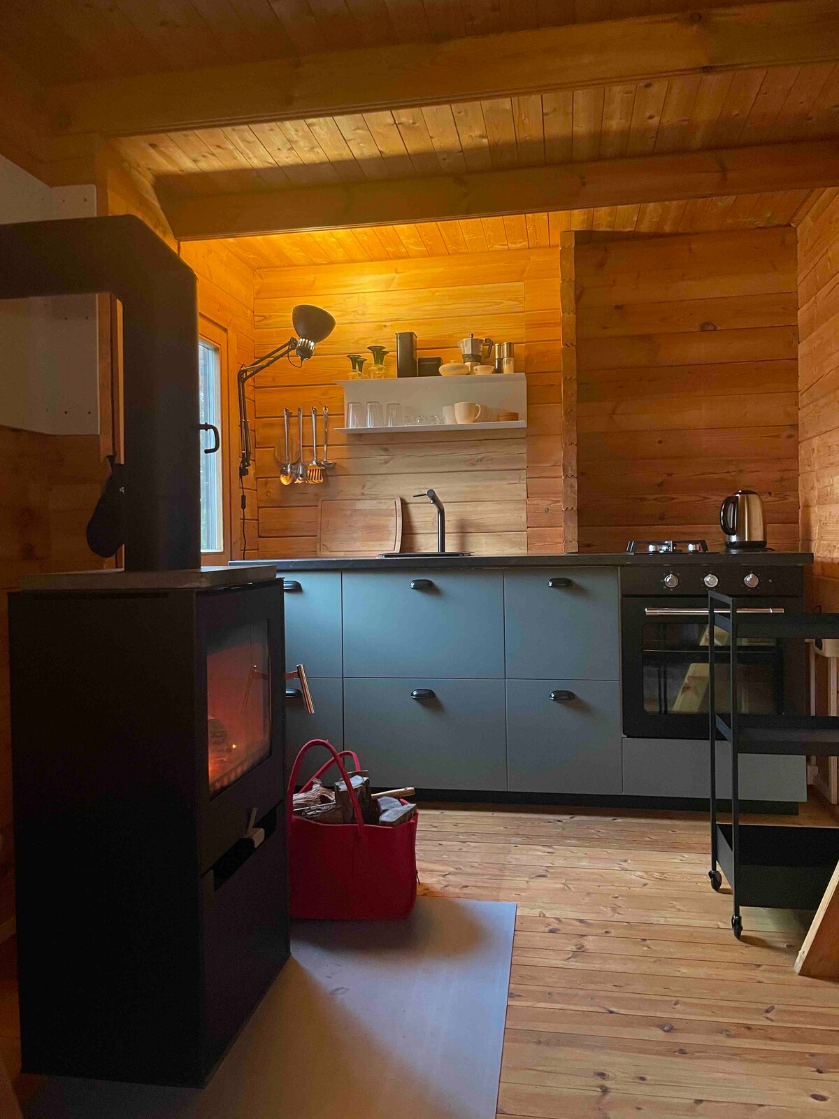 A compact kitchen area features wooden walls and a functional layout. A sleek gas stove, sink, and utensils are provided, along with a modern wood stove. A red bag rests on the floor, complementing the warm, inviting environment illuminated by soft light.