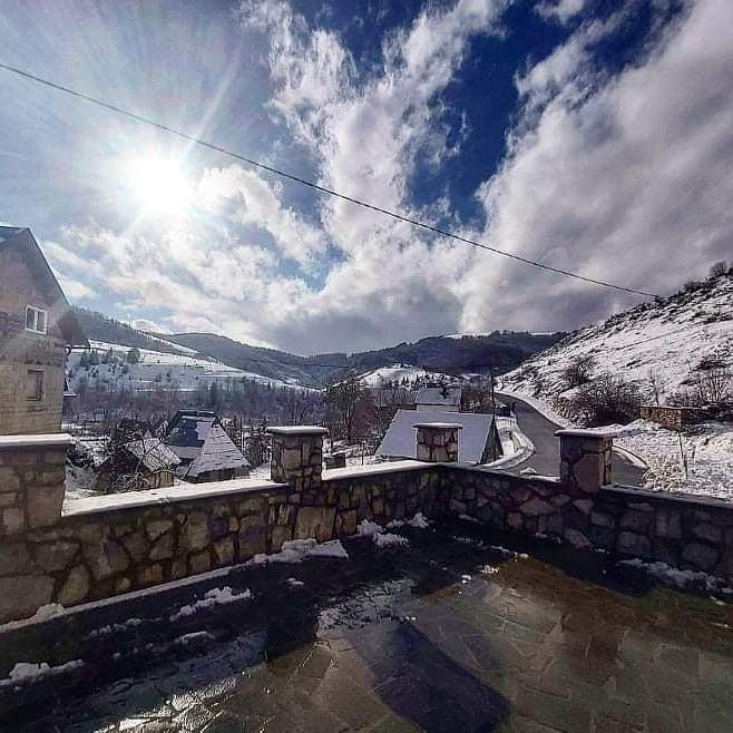 A scenic outdoor view presents rolling hills covered in snow, with sunlight breaking through clouds. Stone walls frame the balcony, while a snowy road meanders through the winter landscape. The tranquil setting showcases nature's beauty in a peaceful winter ambiance.