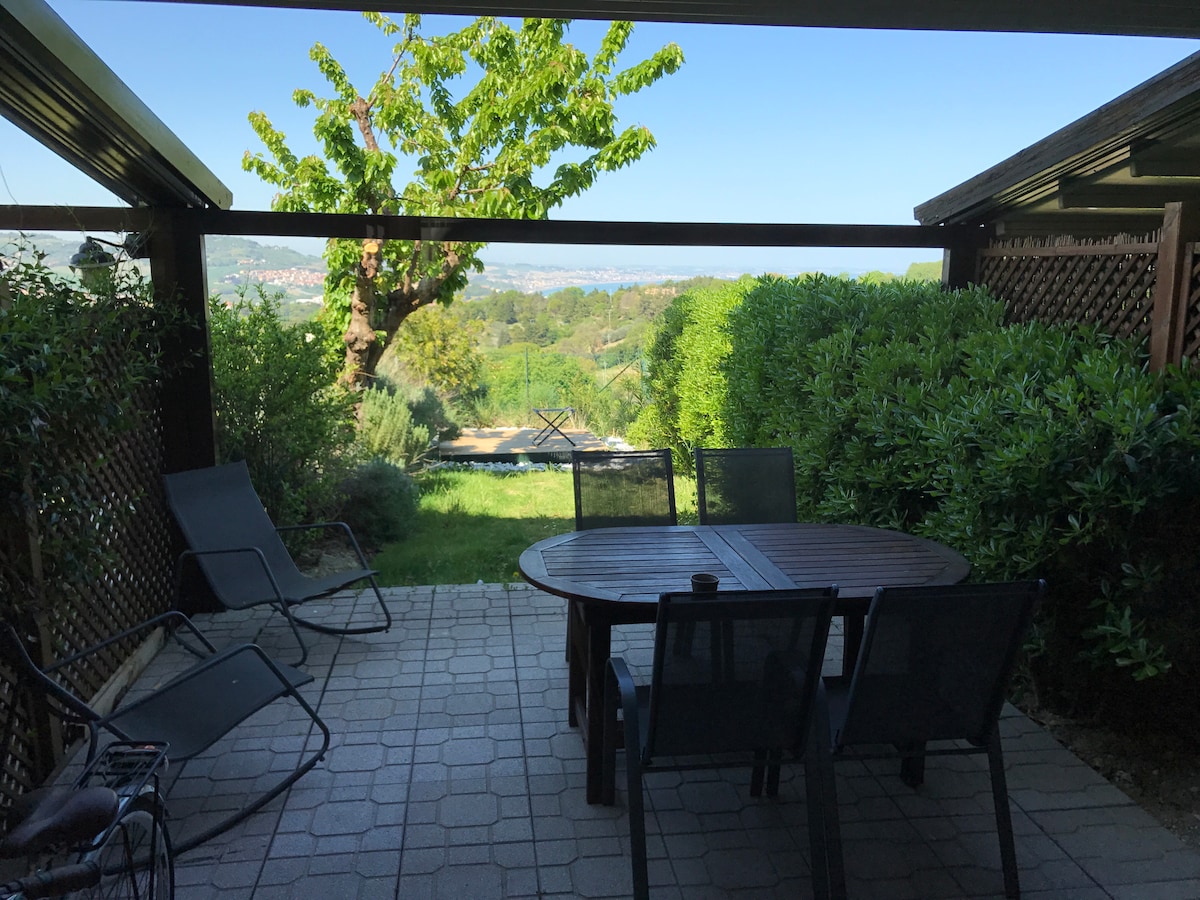 An outdoor patio area features a wooden table surrounded by chairs, with a backdrop of lush greenery and a distant view of rolling hills. Sunlight filters through the leaves of a nearby tree, creating a serene atmosphere ideal for relaxation.