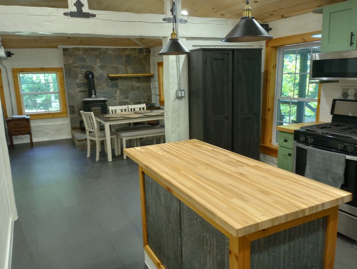 A modern kitchen features a wooden island with a sleek countertop and rustic metal accents. Industrial-style pendant lights hang above. A stone fireplace is visible in the background, alongside a dining table with white chairs. Natural light filters through the windows, highlighting the updated rustic charm.