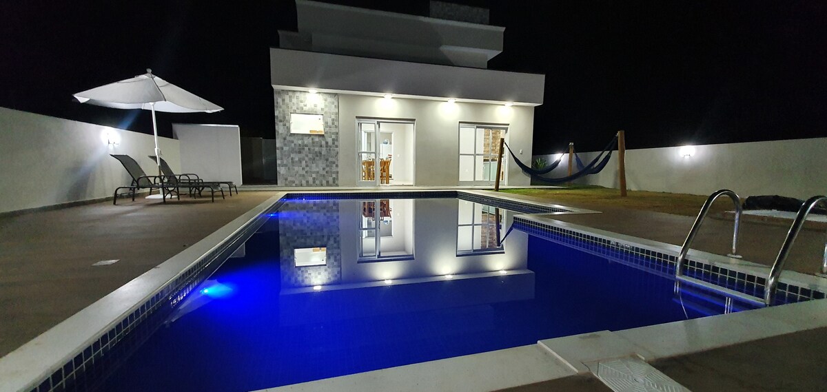 A spacious outdoor area features a modern pool illuminated by subtle lighting, reflecting the house's sleek design. Sun loungers are positioned under an umbrella, and a hammock is visible in the background, creating a serene evening atmosphere.