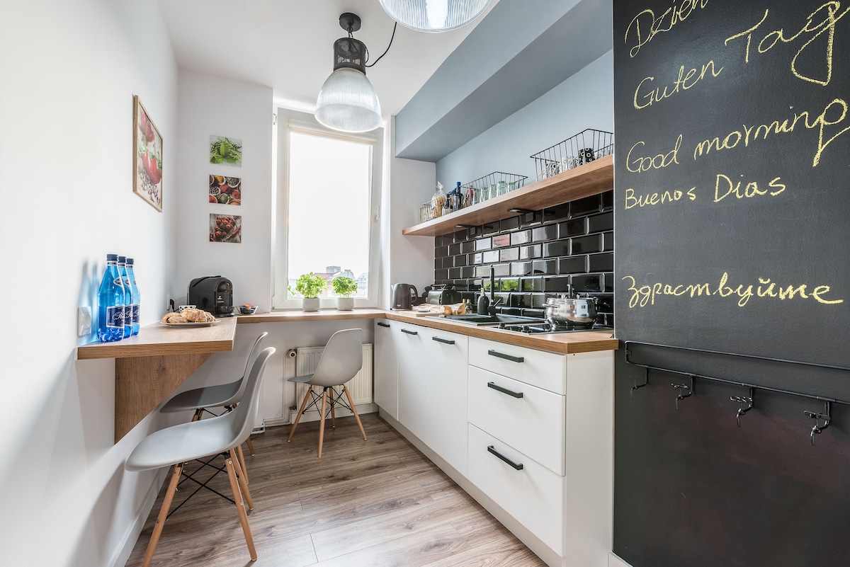 A modern kitchen is presented with light wood and black accents. A wooden table is accompanied by two chairs. The countertop is equipped with a kettle, coffee maker, and kitchen essentials. A chalkboard displays welcoming greetings in multiple languages, and natural light enters through a window.