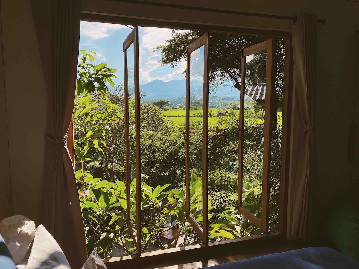 A set of wooden windows is open, revealing a view of lush green paddy fields and distant mountains. Vibrant leaves frame the scene, allowing natural light to fill the interior space, creating a serene and inviting atmosphere.