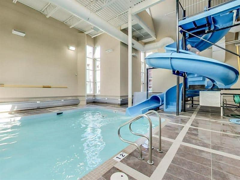 An indoor pool area features a clear blue water pool beside a vibrant blue waterslide. Large windows allow natural light to illuminate the space, creating a welcoming atmosphere. Safety handrails are visible near the pool's edge, ensuring easy access for all guests.
