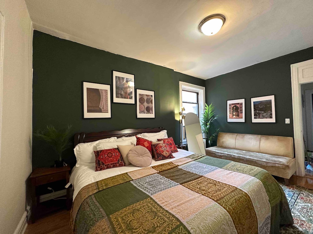 A warm bedroom setting features a spacious bed adorned with a patterned blanket. Photographs and art pieces are displayed on dark green walls. A light source enters through a window. A comfortable sofa and decorative plants complement the inviting atmosphere.