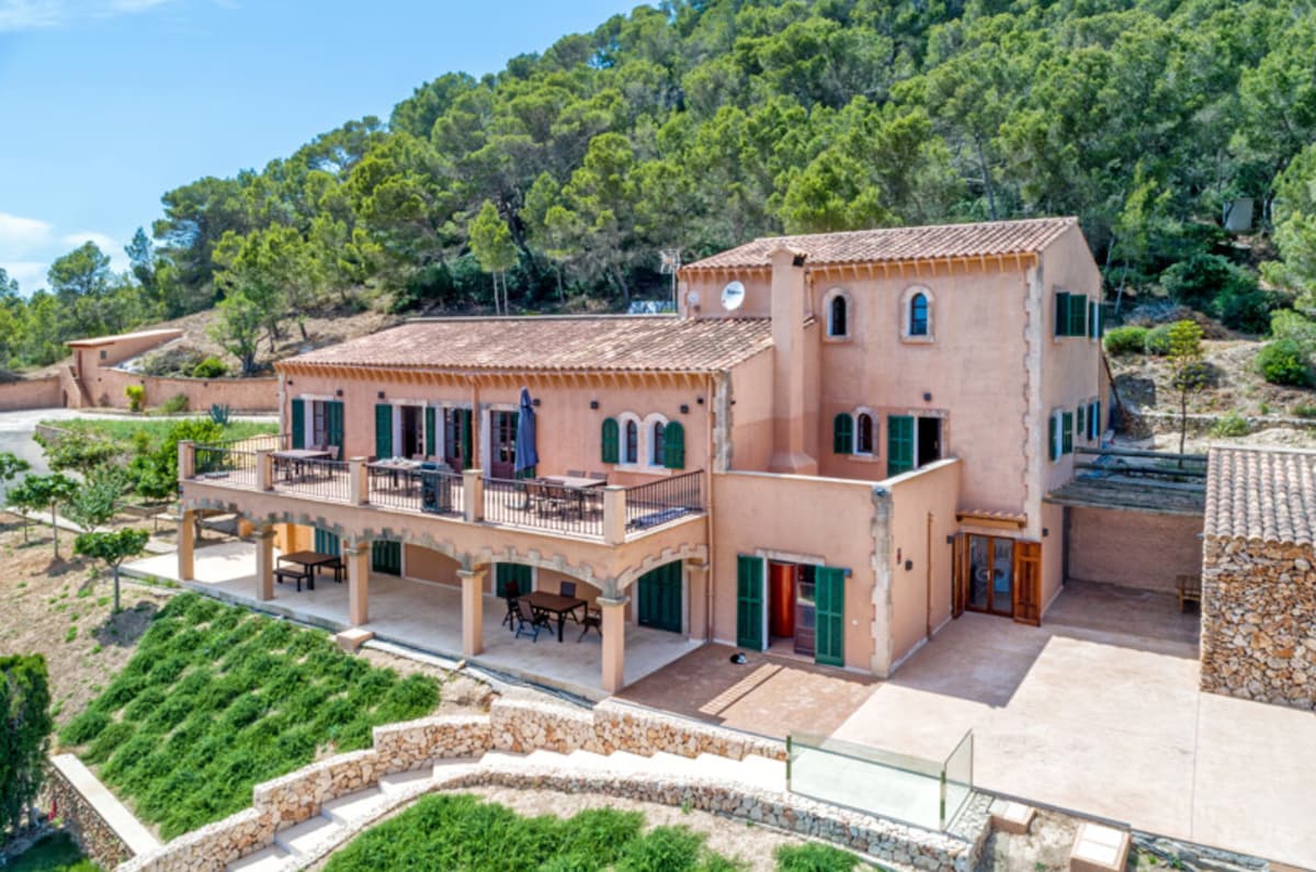 A recently renovated Finca is seen nestled against a hillside, featuring multiple outdoor terraces and an expansive patio. The façade exhibits earthy tones with green shutters. Surrounding greenery and trees create a natural setting, enhancing the property's privacy and peaceful ambiance.