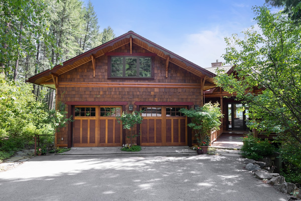 The exterior of a large, rustic home is framed by lush greenery, showcasing a wooden façade with intricate detailing. A spacious driveway leads to two garage doors. Sunlight filters through the trees, creating a serene atmosphere.