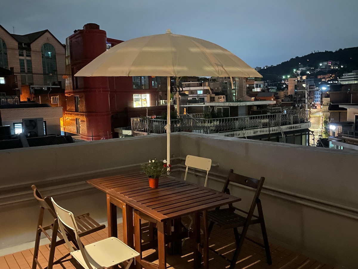 An outdoor terrace is illuminated by soft evening lighting, featuring a wooden table surrounded by white and brown chairs. A large umbrella provides shade, while a potted plant adds a touch of greenery. Urban views showcase surrounding buildings and city streets.