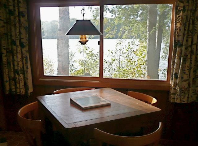 A wooden dining table is set under a pendant light, surrounded by several chairs. A large window offers views of the lake and surrounding trees, bringing natural light into the space.