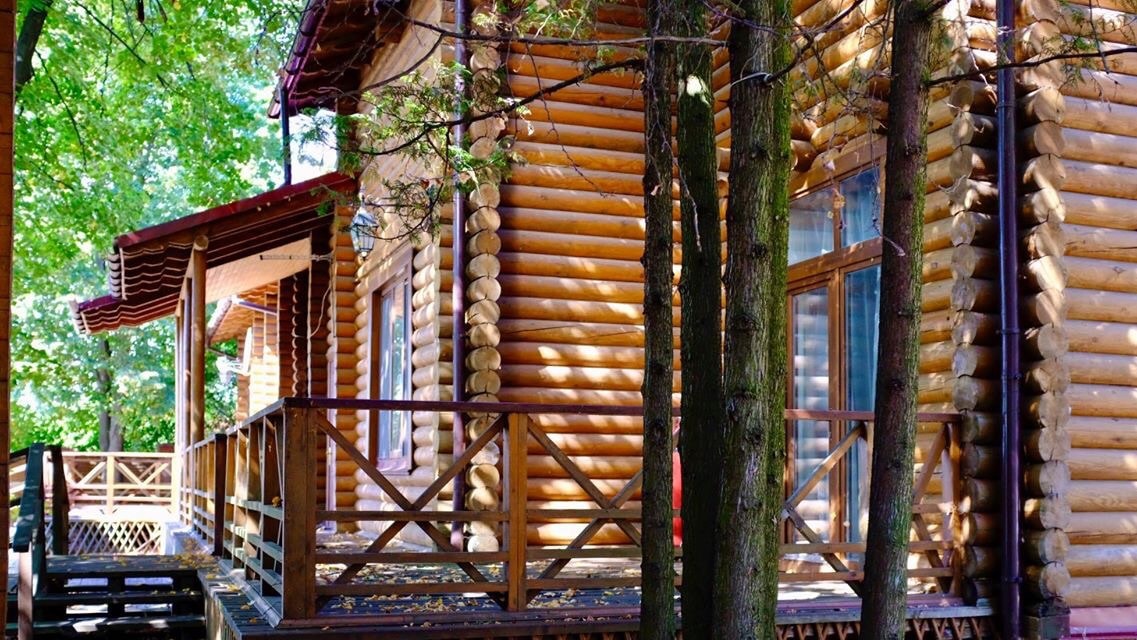 A wooden cabin is surrounded by trees, featuring a covered porch with railings. Large windows are visible, allowing natural light to fill the interior. Sunlight filters through the leaves, creating patterns on the cabin's exterior.