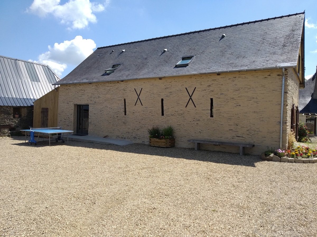 The exterior of a renovated barn is depicted, featuring a light-colored stone façade with decorative cross accents. A spacious gravel courtyard is visible in front, with a wooden bench and colorful flower beds enhancing the outdoor space. Two skylights offer natural light to the interior.