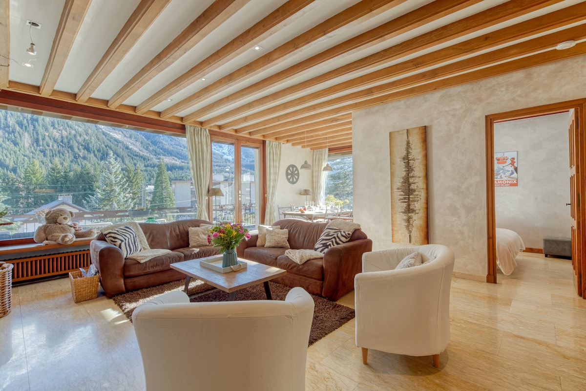 A spacious living area features a comfortable brown sofa facing large windows that provide a view of the surrounding mountains. Natural light fills the space, while a decorative rug and soft seating arrangement create an inviting atmosphere. A stylish coffee table sits at the center.