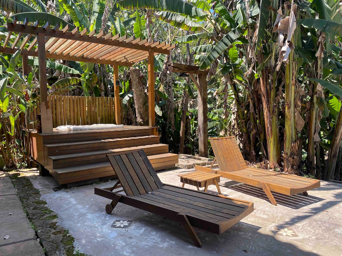 A wooden deck area is surrounded by lush greenery, featuring a shaded seating space with steps leading up to a flat platform. Two wooden lounge chairs are positioned nearby, creating a relaxed atmosphere suitable for outdoor enjoyment.