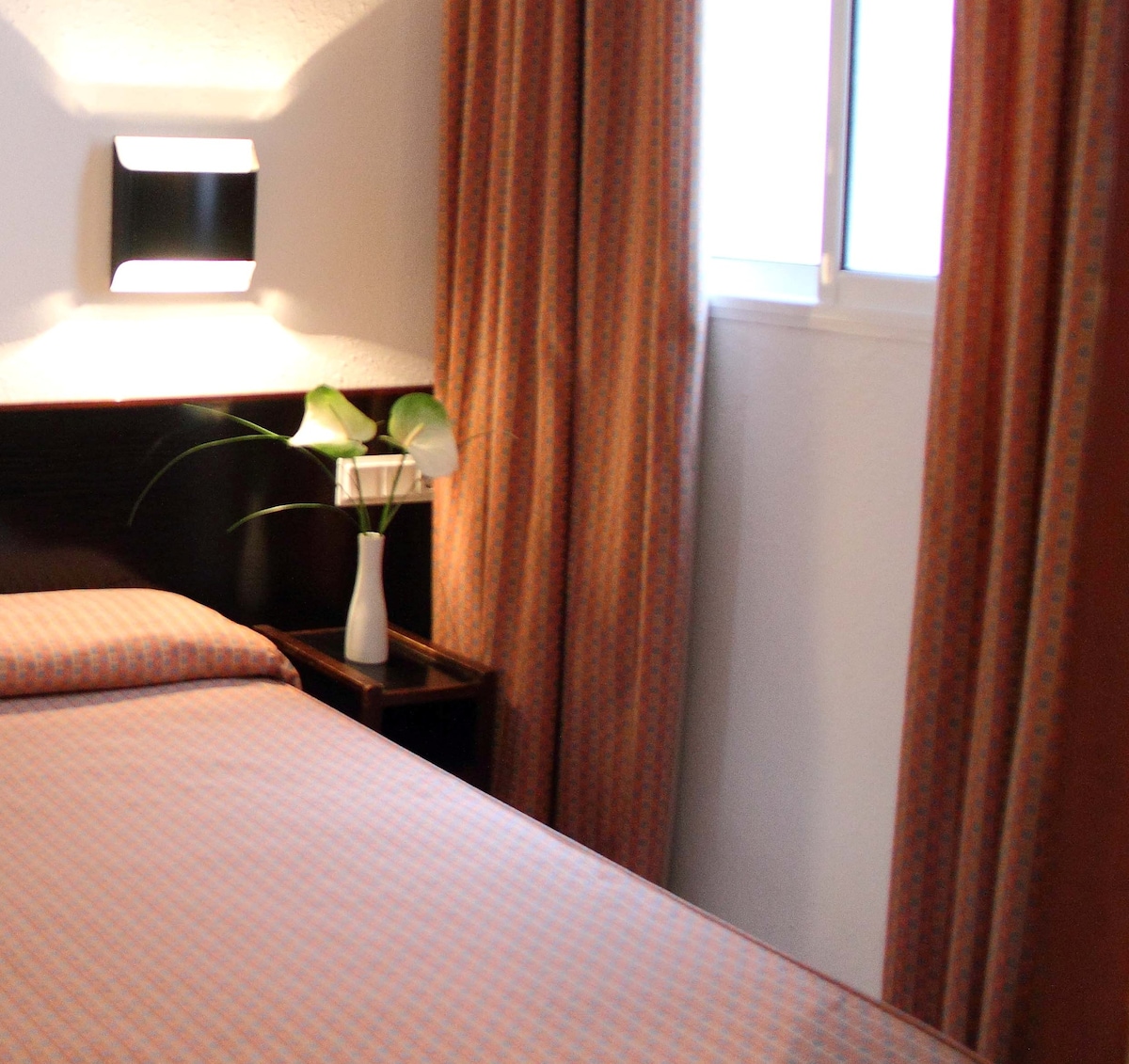 A sleeping area features a spacious bed with a patterned duvet cover, complemented by soft, warm curtains. A small nightstand holds a vase with greenery, and natural light enters through a nearby window.