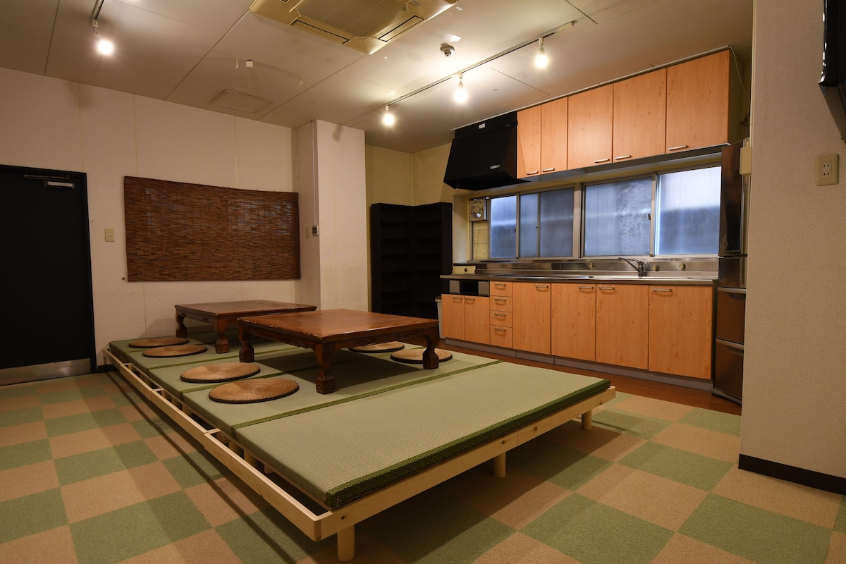 An open space features a traditional low table surrounded by circular cushions on a tatami mat. A modern kitchen with wooden cabinets and stainless-steel appliances is visible in the background, illuminated by soft ceiling lights.