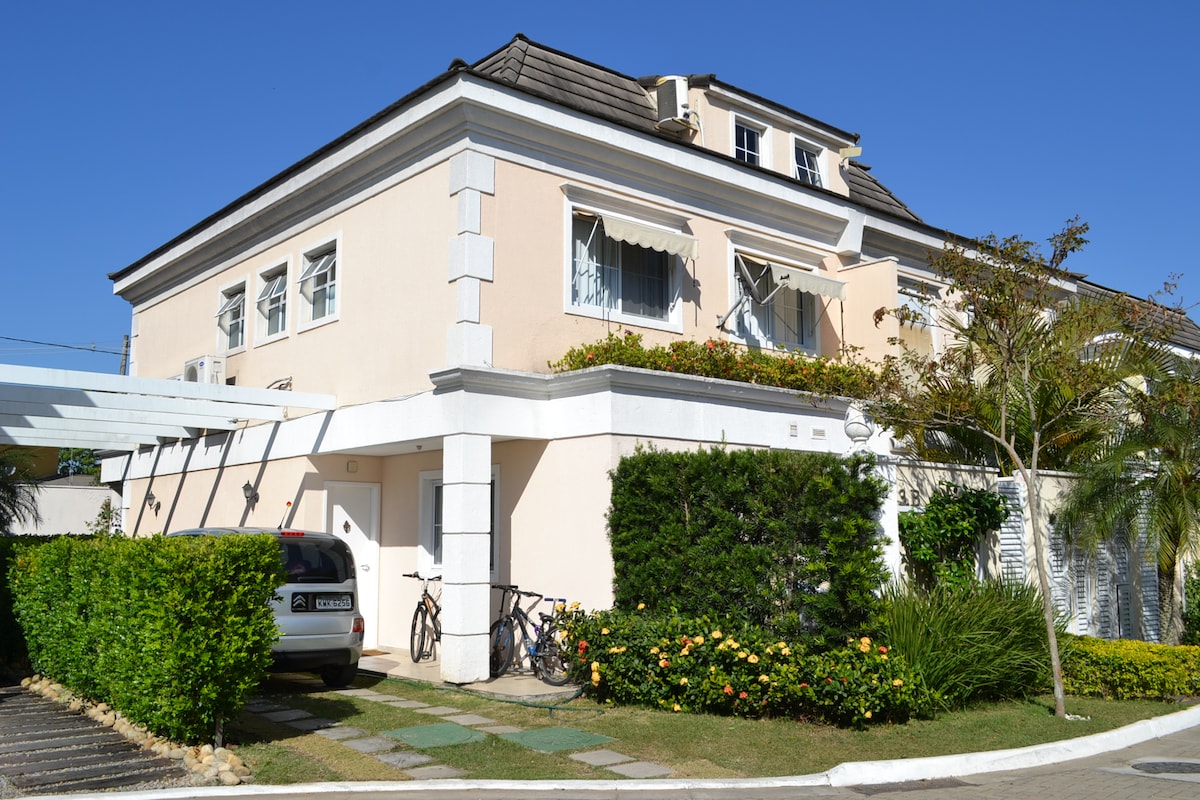 A three-story house is displayed with a light-colored exterior and decorative white trim. Lush greenery and flowering plants surround the entryway, while bicycles are parked in front. A shaded carport accommodates a vehicle beside the well-maintained pathway leading to the entrance.