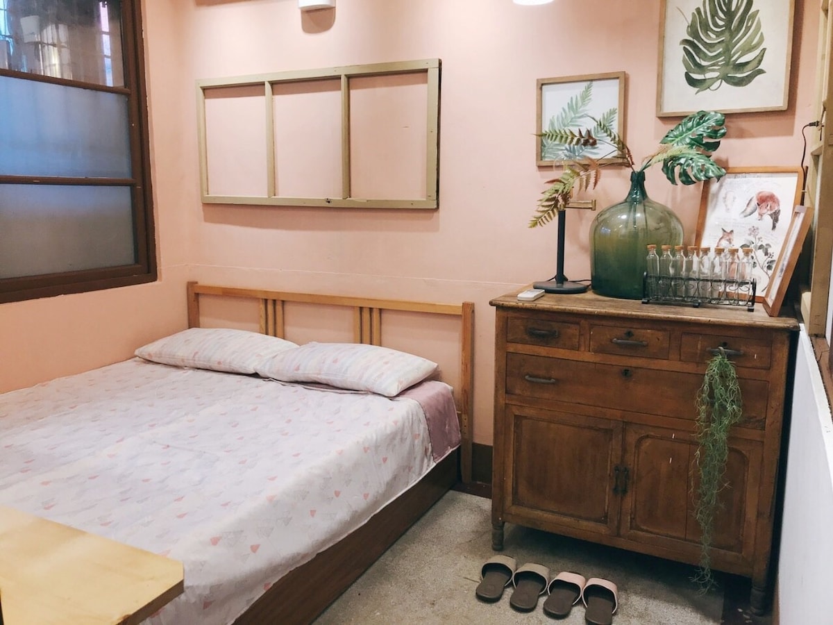 A softly lit sleeping area features a spacious bed with light-colored bedding against a peach-hued wall. A wooden dresser, adorned with decorative plants and framed artwork, complements the warm ambiance. A pair of slippers rests neatly on the floor, adding a touch of home.