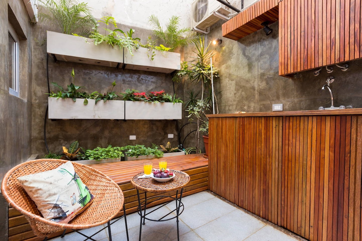 A serene patio space is presented with wooden decking and vertical greenery along the walls. A small table with two chairs is seen, complemented by a beverage setup featuring two glasses and fresh fruit. Natural light illuminates the area, enhancing the inviting environment.