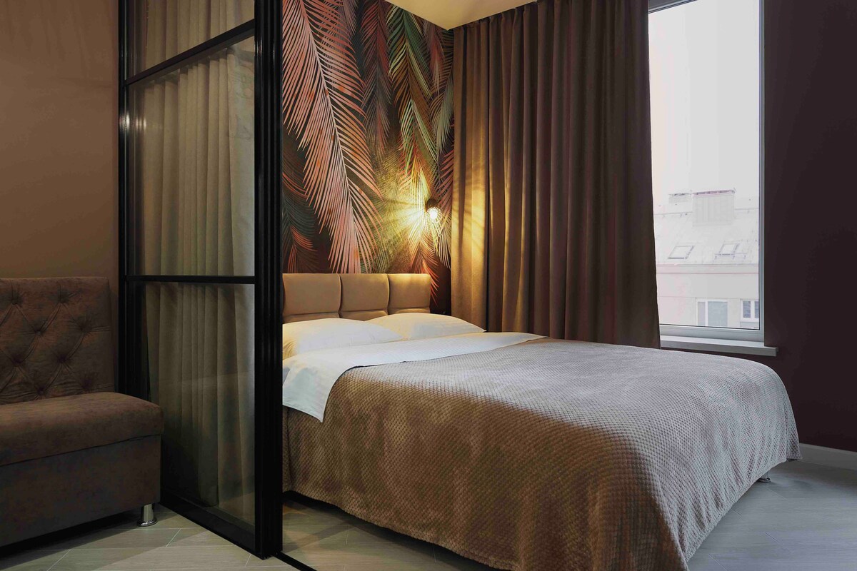A cozy sleeping area features a spacious bed with soft bedding against a backdrop of decorative palm leaf wallpaper. Generous curtains frame a window, allowing natural light to enter. A plush seating area is visible alongside a sleek glass partition.