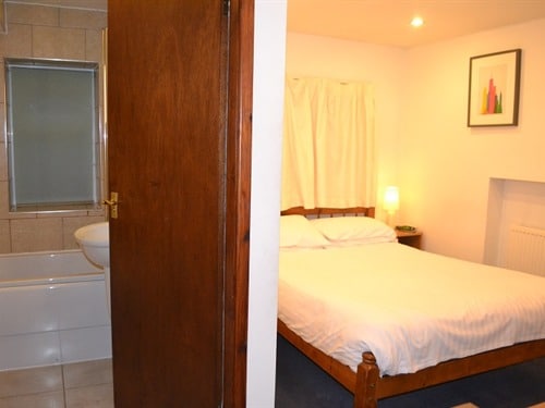 A comfortable room features a spacious bed with crisp white linens. Soft lighting from a bedside lamp enhances the ambiance. A doorway leads to a bathroom, showcasing neutral tiles and a glass shower enclosure.