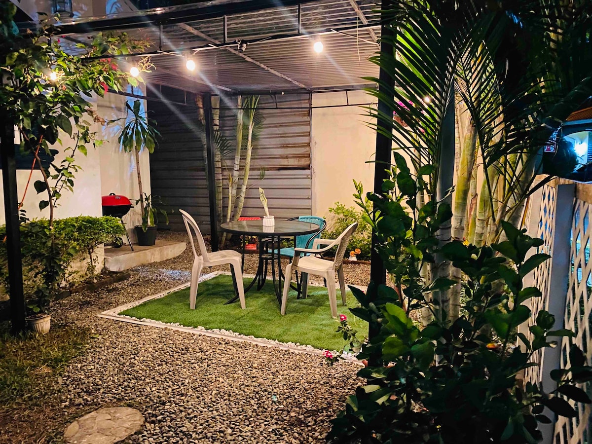 An outdoor garden area is presented, featuring a circular table surrounded by four plastic chairs. Soft lighting illuminates the space, which is bordered by lush green plants and gravel pathways, creating a serene setting for relaxation.