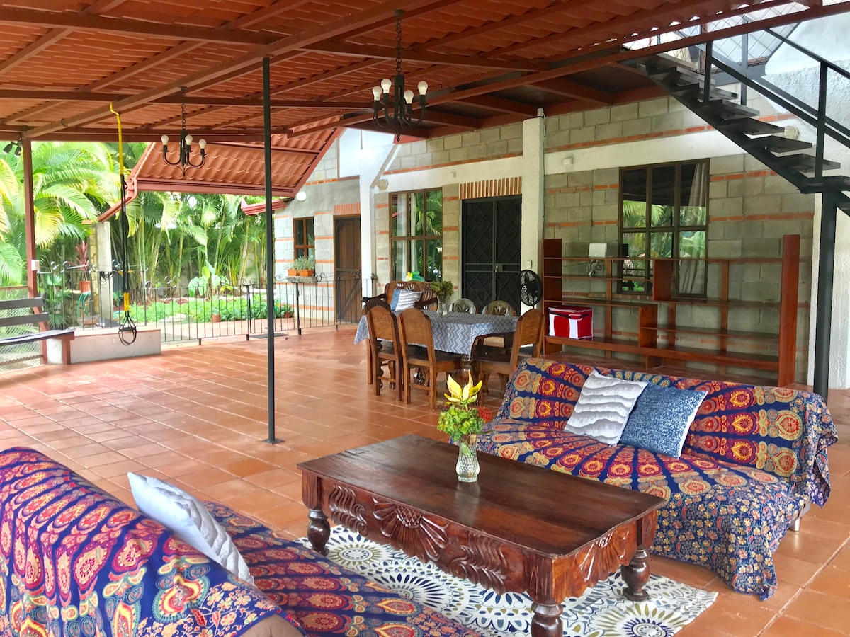 An outdoor living area features a spacious seating arrangement with patterned sofas, a wooden coffee table, and a decorative area rug. Indoor dining tables are visible in the background under a covered terrace, with lush greenery surrounding the space.