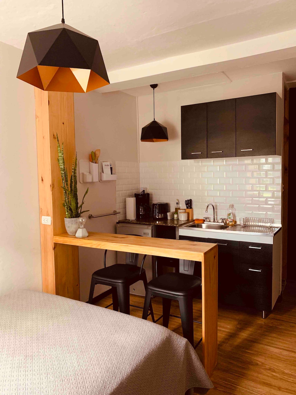 A cozy loft space is shown featuring a kitchen area with modern black cabinets and white tile backsplash. A wooden bar with two black chairs separates the kitchen from the sleeping area, where a neatly made bed is positioned in the foreground.
